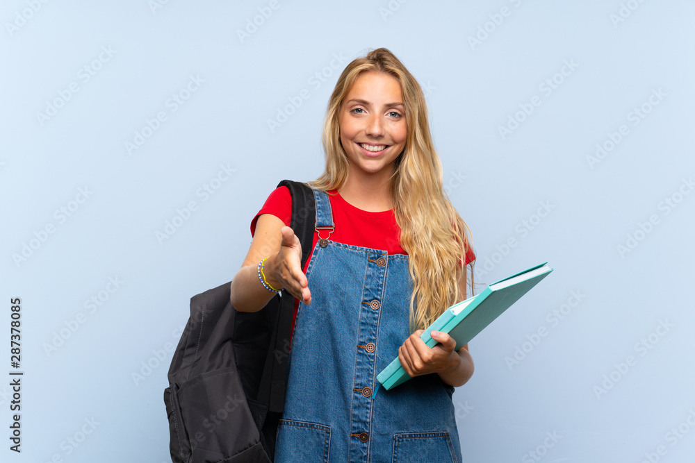 Young blonde student woman over isolated blue wall handshaking after good deal Young blonde student woman over isolated blue wall handshaking after good deal