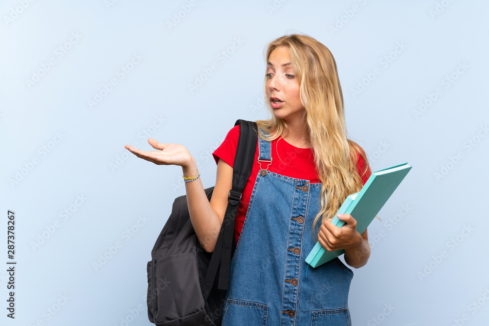 Young blonde student woman over isolated blue wall holding copyspace imaginary on the palm Young blonde student woman over isolated blue wall holding copyspace imaginary on the palm