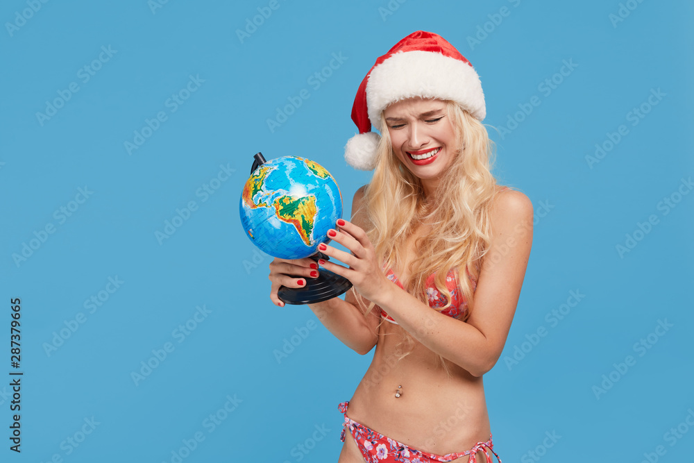 Suntan crying and upset young woman with slim body in santa hat and bikini holding a globe. Christmas sunny day summer vacation and holiday concept. Not enough money for a ticket Suntan crying and upset young woman with slim body in santa hat and bikini holding a globe. Christmas sunny day summer vacation and holiday concept. Not enough money for a ticket