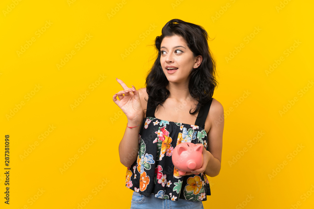 Young woman over isolated yellow background holding a big piggybank Young woman over isolated yellow background holding a big piggybank