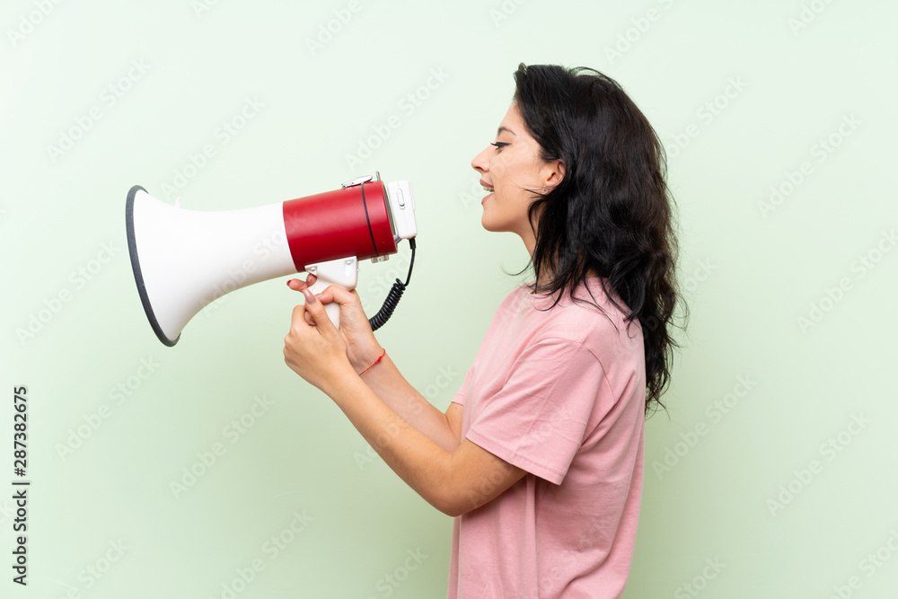 Young woman over isolated green background shouting through a megaphone Young woman over isolated green background shouting through a megaphone