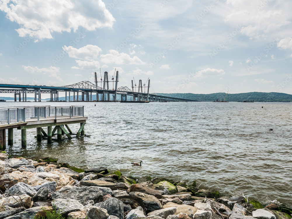 The Tappan Zee Bridge under construction in New York with cranes as seen from the shoreline with rocks along the coast.