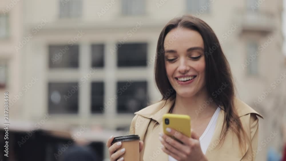 Young Pretty Lady Looking Happy Drinking Coffe Smiling Using her Smartphone Impressed Saying Wow Walking at City Street Close Up. Young Pretty Lady Looking Happy Drinking Coffe Smiling Using her Smartphone Impressed Saying Wow Walking at City Street Close Up.