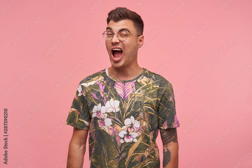 Cheerful young pretty male in glasses looking at camera and giving wink with broad smile, wearing glasses and flowered t-shirt, isolated over pink background Cheerful young pretty male in glasses looking at camera and giving wink with broad smile, wearing glasses and flowered t-shirt, isolated over pink background