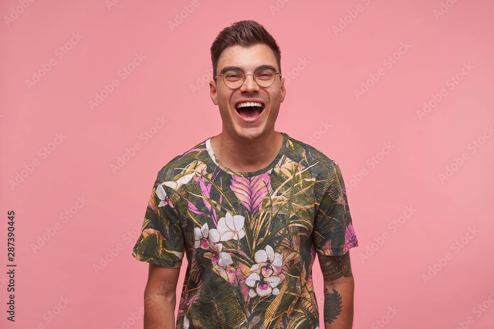 Close-up of joyful attractive young male with short haircut standing over pink background with hands down, being in nice mood, laughing at funny joke Close-up of joyful attractive young male with short haircut standing over pink background with hands down, being in nice mood, laughing at funny joke