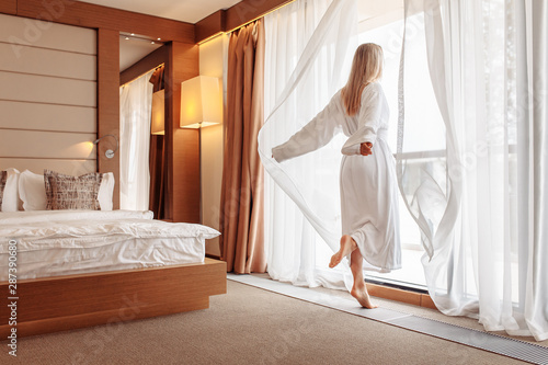Joyful emotional young beautiful woman in white bathrobe happily opens the curtains of her window in the bedroom on a sunny summer morning. The concept of starting a new day and fulfilling your plans
