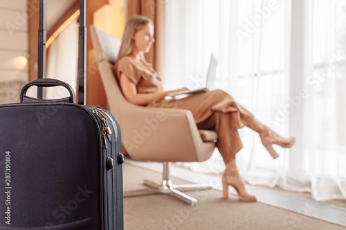 Suitcase on backdrop of blurred young woman traveler looking for best room into hotel while traveling. Businesswoman leafing through online catalog of hotel spa services sitting in comfortable chair
