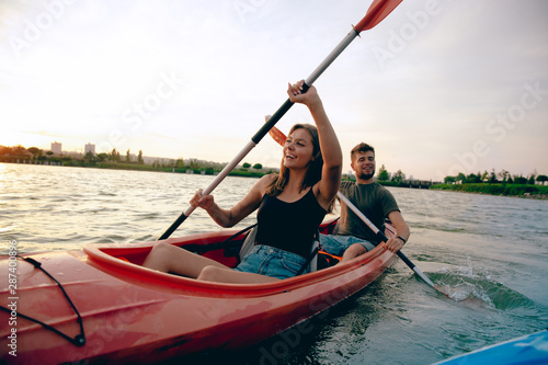 Confident young caucasian couple kayaking on river together with sunset in the backgrounds. Having fun in leisure activity. Romantic and happy woman and man on the kayak. Sport, relations concept. © master1305