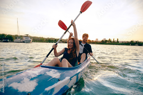 Confident young caucasian couple kayaking on river together with sunset in the backgrounds. Having fun in leisure activity. Romantic and happy woman and man on the kayak. Sport, relations concept.
