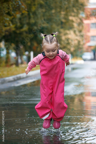 Happy child girl with pink rubber pants and boots in puddle on an autumn walk, seasonal fun childhood game photo