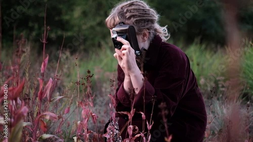 Trendy hipster girl with dreads taking pictures of plants in the garden with a vintage film camera at cold summer morning photo