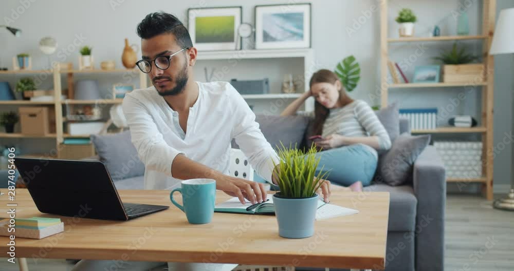 Man freelancer is working with laptop at home then writing in notebook while girl is using smartphone on sofa in modern apartment. Youth and lifestyle concept. Man freelancer is working with laptop at home then writing in notebook while girl is using smartphone on sofa in modern apartment. Youth and lifestyle concept.