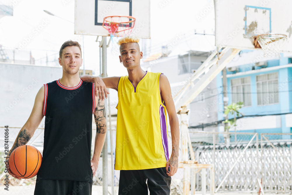 Smiling young multi-ethnic basketball players in uniform standing at street court and looking at camera Smiling young multi-ethnic basketball players in uniform standing at street court and looking at camera