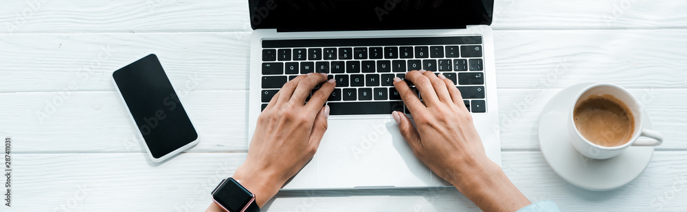 panoramic shot of woman typing on laptop near smartphone and cup of coffee