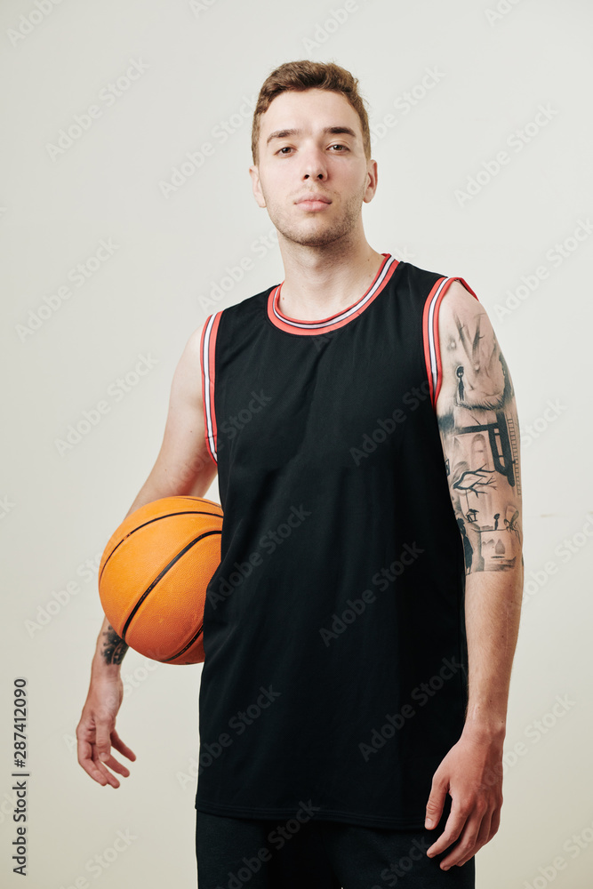 Confident serious backetball player in black uniform holding ball and looking at camera, isolated on white