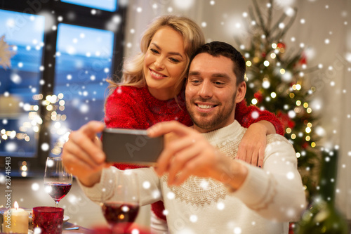 christmas, holidays, technology and people concept - happy couple in taking selfie by smartphone at home dinner over snow
