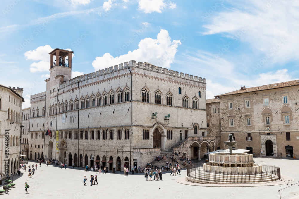 Photograph of the main square of Perugia (Piazza 4 Novembre), with a view of the Fontana Maggiore and the town hall. In the background blue sky and clouds.