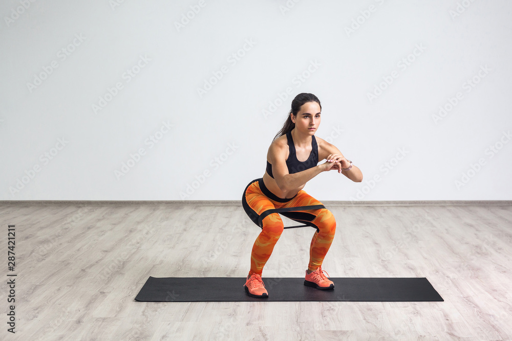 Portrait of young sporty healthy beautiful woman in black top and orange leggings doing squatting with elastic resistance band. Isolated, white wall, indoor, workout concept, looking away