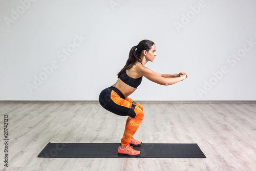 Fotografie Side view portrait of young sporty healthy beautiful woman in black top and orange leggings doing squatting with elastic resistance band