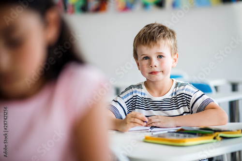 A small school boy sitting at the desk in classroom  looking at camera.