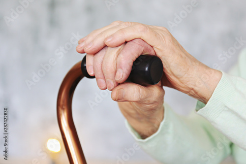 Elderly woman sitting in nursing home room holding walking quad cane with wrinked hand. Old age senior lady wearing beige cardigan, metal aid stick handle bar close up. Interior background, copy space photo