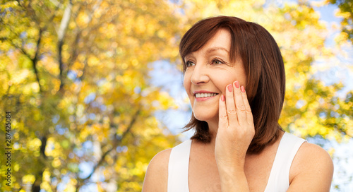 beauty, skin care and old people concept - portrait of smiling senior woman touching her face over natural autumn background