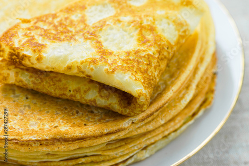 Stack of folded delicious freshly baked crepes with appetizing golden crust on white plate on linen table cloth by window in sunlight. Cozy atmosphere breakfast morning
