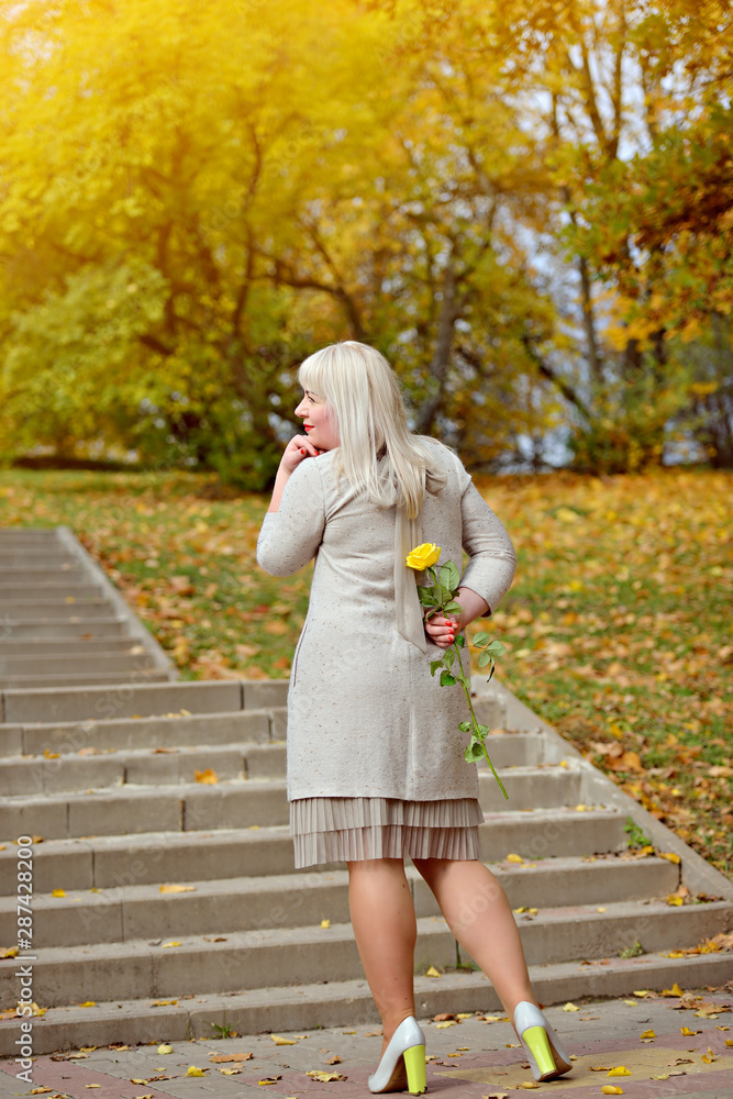Cute blonde big size stands and holds a yellow rose behind his back on a Sunny autumn day in the Park, against a stone staircase. She looks away thoughtfully. Conceptual photography. Rear view Cute blonde big size stands and holds a yellow rose behind his back on a Sunny autumn day in the Park, against a stone staircase. She looks away thoughtfully. Conceptual photography. Rear view