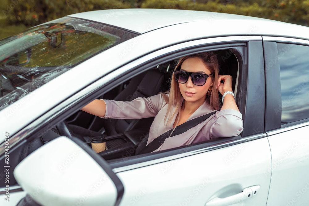 Beautiful woman driving a car, in the summer in the city, sunglasses, pink suit, tanned figure, close-up portrait, white business class sedan, city parking, business woman.