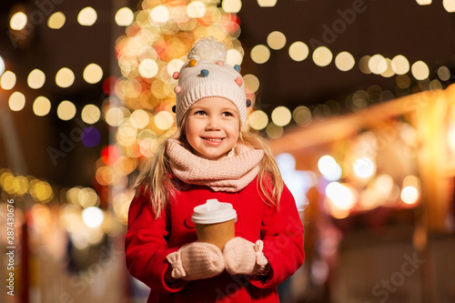 holidays, childhood and people concept - happy little girl with cup of tea at christmas market in winter evening