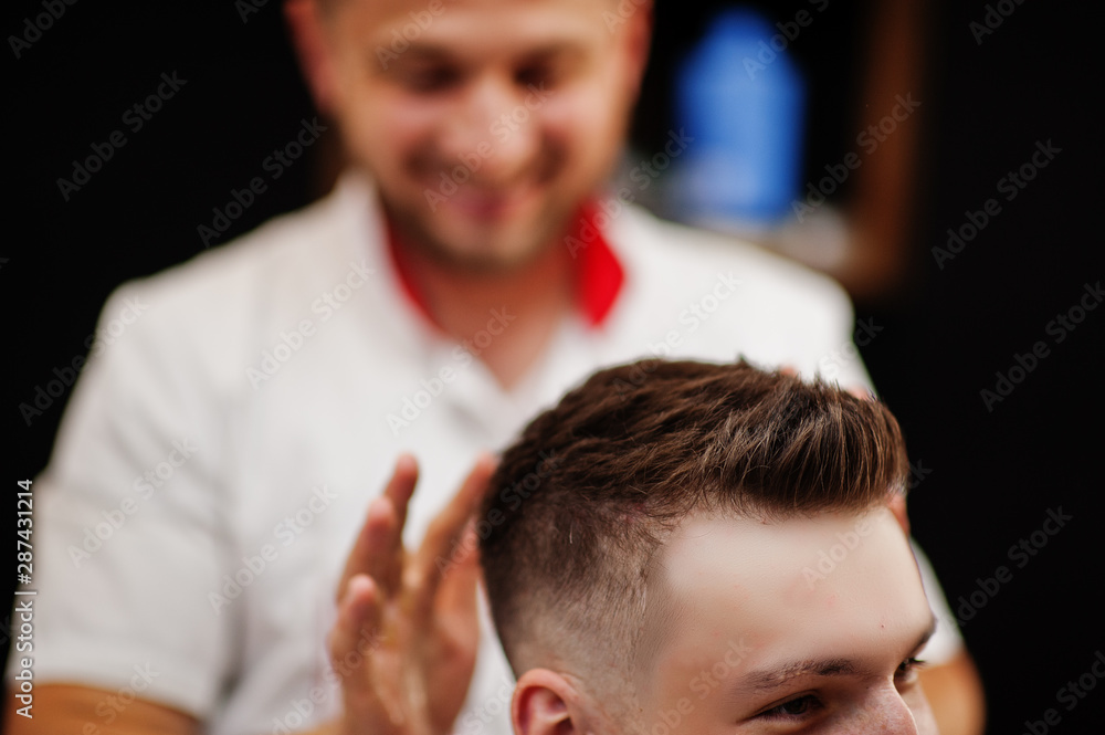 Fototapeta premium Young bearded man getting haircut by hairdresser while sitting in chair at barbershop. Barber soul.