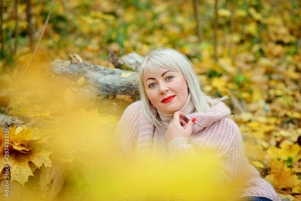 Naklejka premium A bright middle-aged blonde poses on a Sunny autumn day in the forest in a gentle knitted jumper. Photo through the foliage. Woman's size plus xxl. Horizontal view