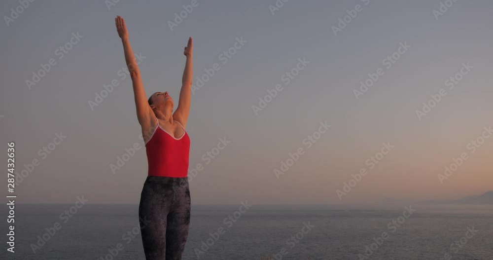 Caucasian woman in red relaxing by practicing yoga on the cliff near calm sea at sunset. Slow motion Caucasian woman in red relaxing by practicing yoga on the cliff near calm sea at sunset. Slow motion
