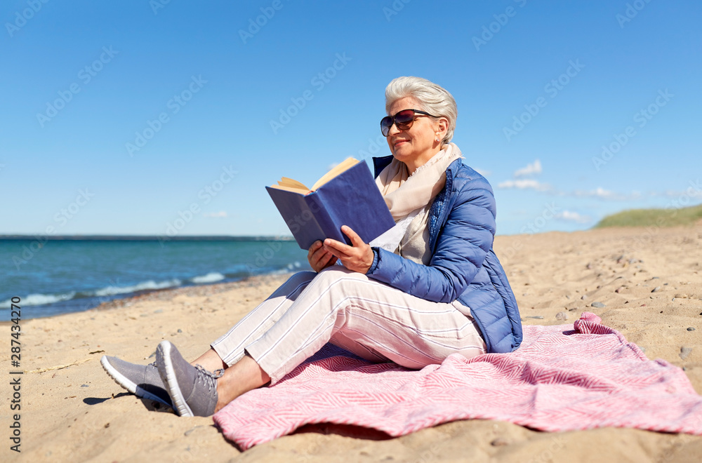 people and leisure concept - happy senior woman reading book on summer beach in estonia