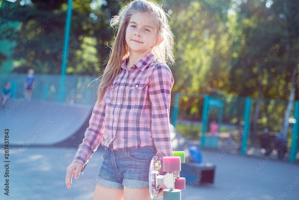 Naklejka premium portrait of a beautiful little teenage girl, holding a skateboard, against the backdrop of a beautiful sun, in skatepark