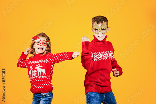 Confident children in warm red Christmas sweaters and decorated glasses looking at camera on yellow background