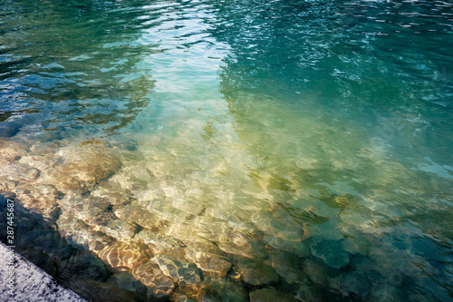 Lake or pond surface. Stone bed is seen through transparent water. Green and blue tints with sunlight flecks on it