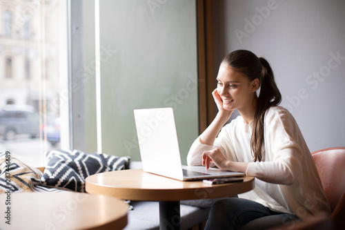 Young smiling hipster girl talking with friend via video call via laptop computer while sitting in coffee shop. Cheerful female manager having webinar via portable notebook during resting time in cafe
