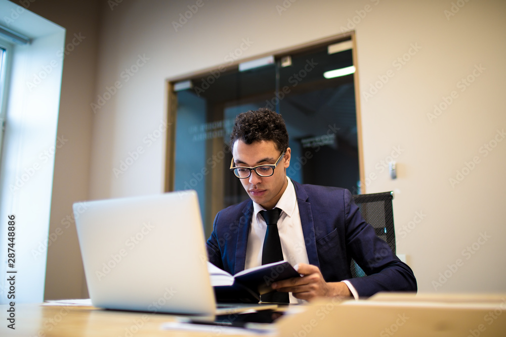 Professional Afro businessman in formal suit reading information in textbook, sitting at table with open laptop computer. Male entrepreneur preparing to meeting with staff, using netbook and notebook Professional Afro businessman in formal suit reading information in textbook, sitting at table with open laptop computer. Male entrepreneur preparing to meeting with staff, using netbook and notebook