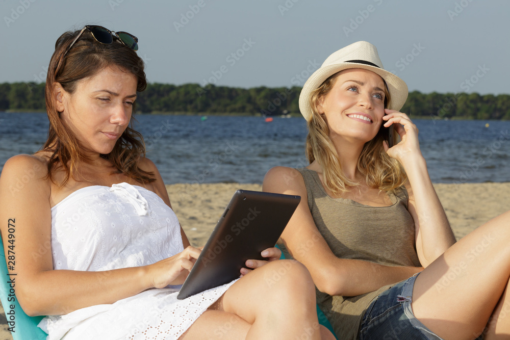 two women using technology while relaxing on the beach