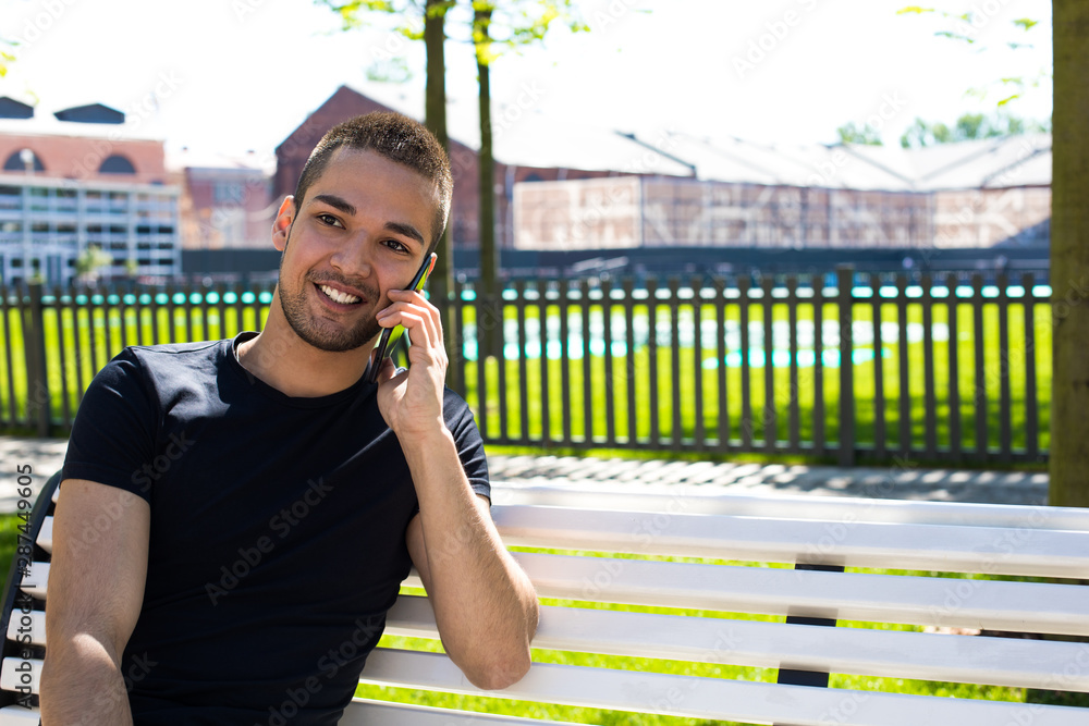 Fototapeta premium Young smiling hipster guy talking with friend via mobile phone while sitting on the bench in city park in beautiful sunny summer day. Cheerful man having pleasant cell telephone conversation