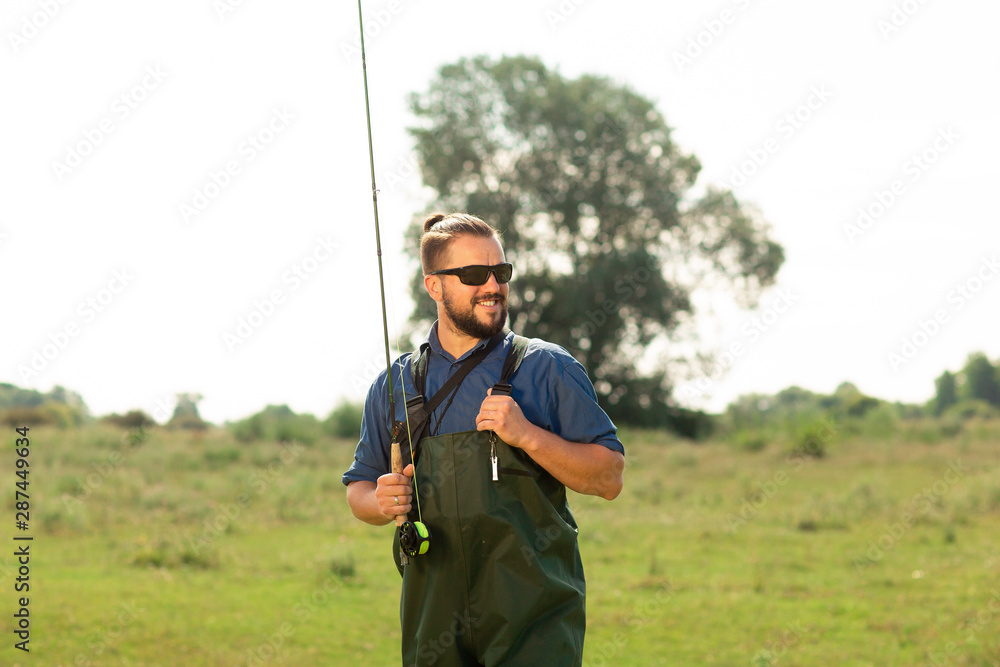 Happy fisherman in black glasses and a special suit poses in the nature. He holds his fishing rod in his hands - obrazy, fototapety, plakaty 