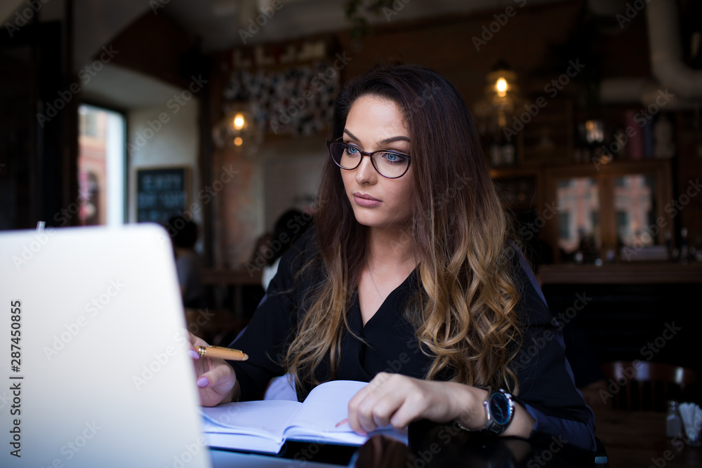 Female wearing in glasses smart college student using textbook during online education via laptop computer while sitting in cozy coffee shop interior. Woman skilled blogger having webinar via netbook