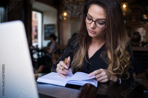 Woman with long hair wearing in stylish glasses writing notary in textbook during online learning on laptop computer, sitting in restaurant interior. Female marketing coordinator using diary for work