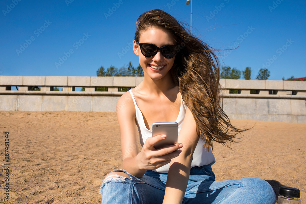 Beautiful smiling woman in fashionable sunglasses reading pleasant text message in social network via mobile phone, sitting on the beach in sunny summer day. Happy female watching video on smartphone