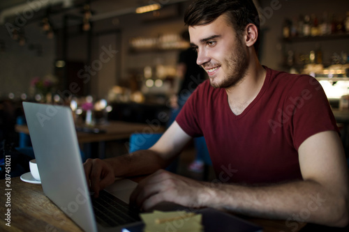 Young smiling man university student having online education via laptop computer while sitting in modern coffee shop. Joyful handsome male chatting via notebook during recreation time in restaurant © ZoFot