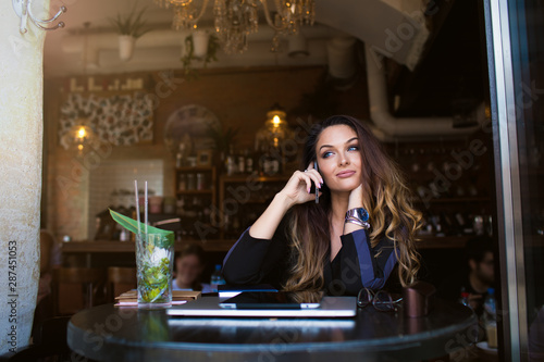 Young woman with long beautiful hair talking via mobile phone during recreation time in coffee shop, sitting near copy space for promotional content. Female university student having smartphone call