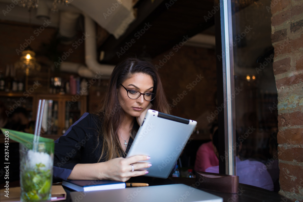Woman wearing in glasses reading e-book on digital tablet during rest in coffee shop. Attractive thoughtful female online booking via touch pad gadget. Women having training course Woman wearing in glasses reading e-book on digital tablet during rest in coffee shop. Attractive thoughtful female online booking via touch pad gadget. Women having training course