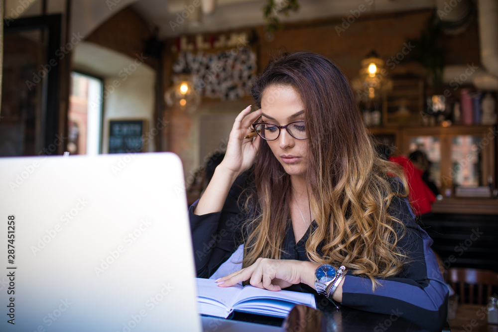 Woman professional writer reading textbook during work on laptop computer while sitting in coffee shop. Female university student wearing in stylish glasses learning with notepad and net-book. Woman professional writer reading textbook during work on laptop computer while sitting in coffee shop. Female university student wearing in stylish glasses learning with notepad and net-book.
