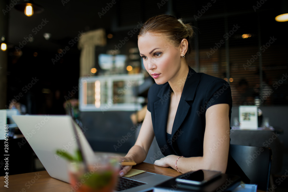 Serious woman financier reading information on stock exchange via modern laptop computer while sitting in restaurant during work break in enterprise. Female jurist having video conference via netbook Serious woman financier reading information on stock exchange via modern laptop computer while sitting in restaurant during work break in enterprise. Female jurist having video conference via netbook